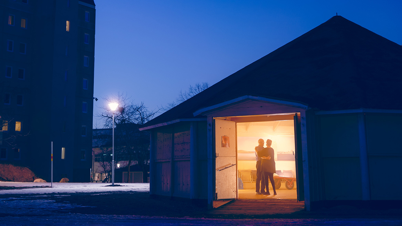 Ett bostadsområde i skymningen, en dörr är öppen till en lokal där det lyser varmt på ett dansande par.
